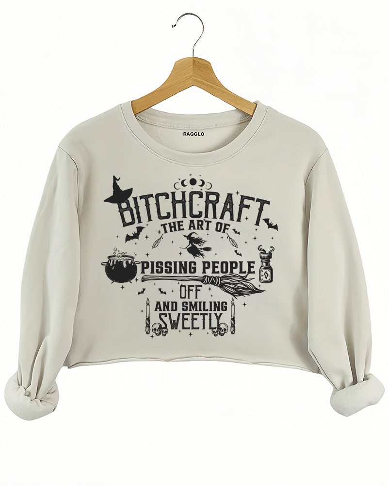 Bitchcraft design featuring the phrase 'The Art of Pissing People Off and Smiling Sweetly' along with witchcraft elements like a cauldron, broom, and skull candles, printed on a beige crop sweatshirt available at Ragglo.