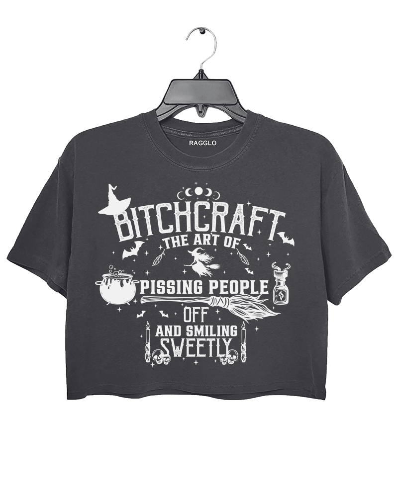 Bitchcraft graphic featuring the phrase 'The Art of Pissing People Off and Smiling Sweetly,' with witchcraft elements like a cauldron, broom, flying witch, and magic potion, printed on a dark gray crop top - available on Ragglo.