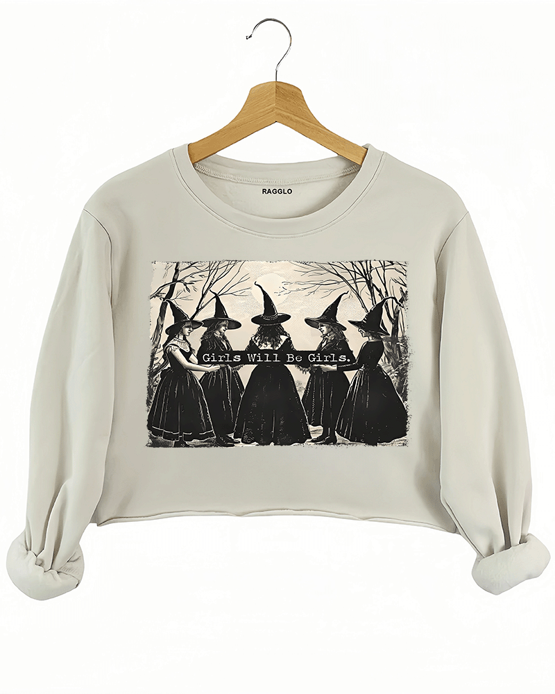 Ragglo beige women's crop sweatshirt featuring five witches in black hats holding a sign with the text 'Girls Will Be Girls' in a spooky forest background.
