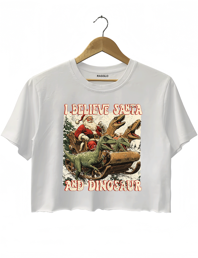 Ragglo white women's crop top with 'I Believe Santa and Dinosaur' text and Santa sleigh pulled by dinosaurs graphic.