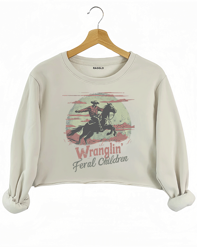 Ragglo beige women's crop sweatshirt featuring a cowboy riding a horse under a moonlit mountain background with the humorous text 'Wranglin' Feral Children' in a playful western graphic design.