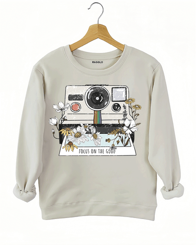 Ragglo beige sweatshirt featuring a vintage Polaroid camera surrounded by flowers, with 'FOCUS ON THE GOOD' text, perfect for retro-inspired streetwear with a positive vibe.