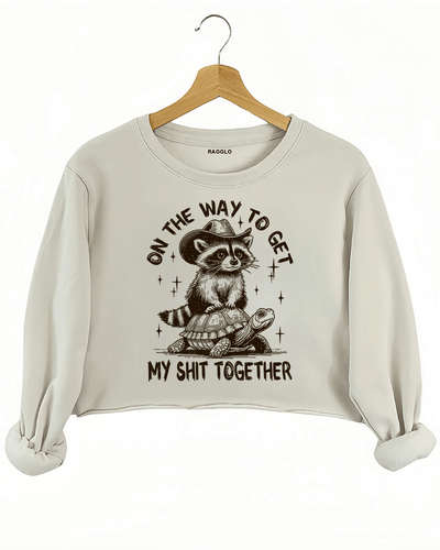 Ragglo beige crop sweatshirt featuring 'ON THE WAY TO GET MY SHIT TOGETHER' text with a raccoon in a cowboy hat riding a turtle, perfect for casual streetwear and humorous everyday wear.