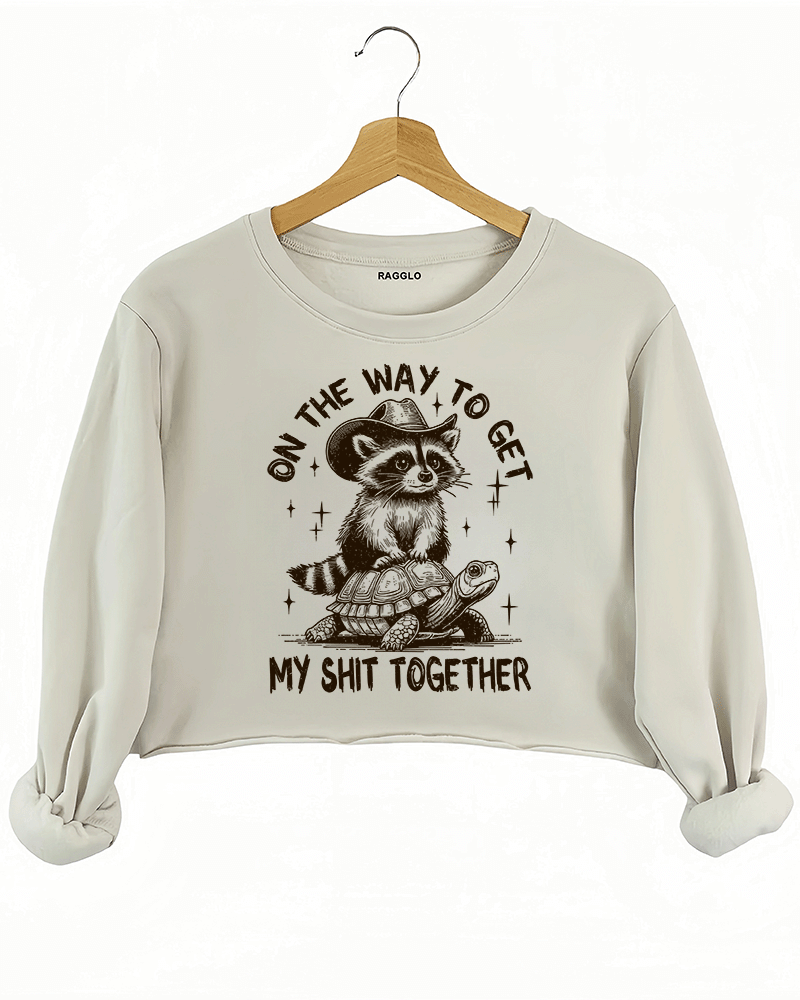 Ragglo beige crop sweatshirt featuring 'ON THE WAY TO GET MY SHIT TOGETHER' text with a raccoon in a cowboy hat riding a turtle, perfect for casual streetwear and humorous everyday wear.