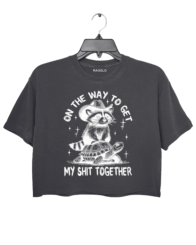 Ragglo dark gray crop top featuring 'ON THE WAY TO GET MY SHIT TOGETHER' text with a raccoon in a cowboy hat riding a turtle, perfect for casual streetwear and humorous everyday wear.