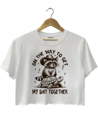 Ragglo white crop top featuring 'ON THE WAY TO GET MY SHIT TOGETHER' text with a raccoon in a cowboy hat riding a turtle, perfect for casual streetwear and humorous everyday wear.