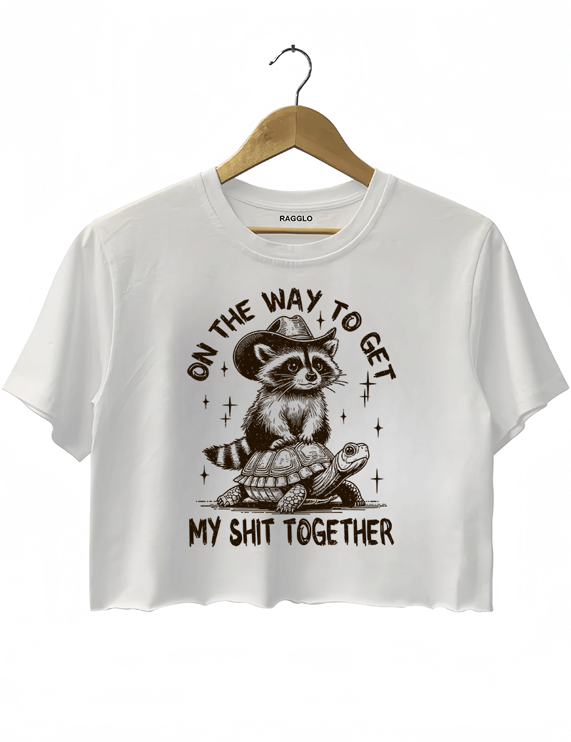 Ragglo white crop top featuring 'ON THE WAY TO GET MY SHIT TOGETHER' text with a raccoon in a cowboy hat riding a turtle, perfect for casual streetwear and humorous everyday wear.