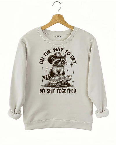 Ragglo beige sweatshirt featuring 'ON THE WAY TO GET MY SHIT TOGETHER' text with a raccoon in a cowboy hat riding a turtle, perfect for casual streetwear and humorous everyday wear.