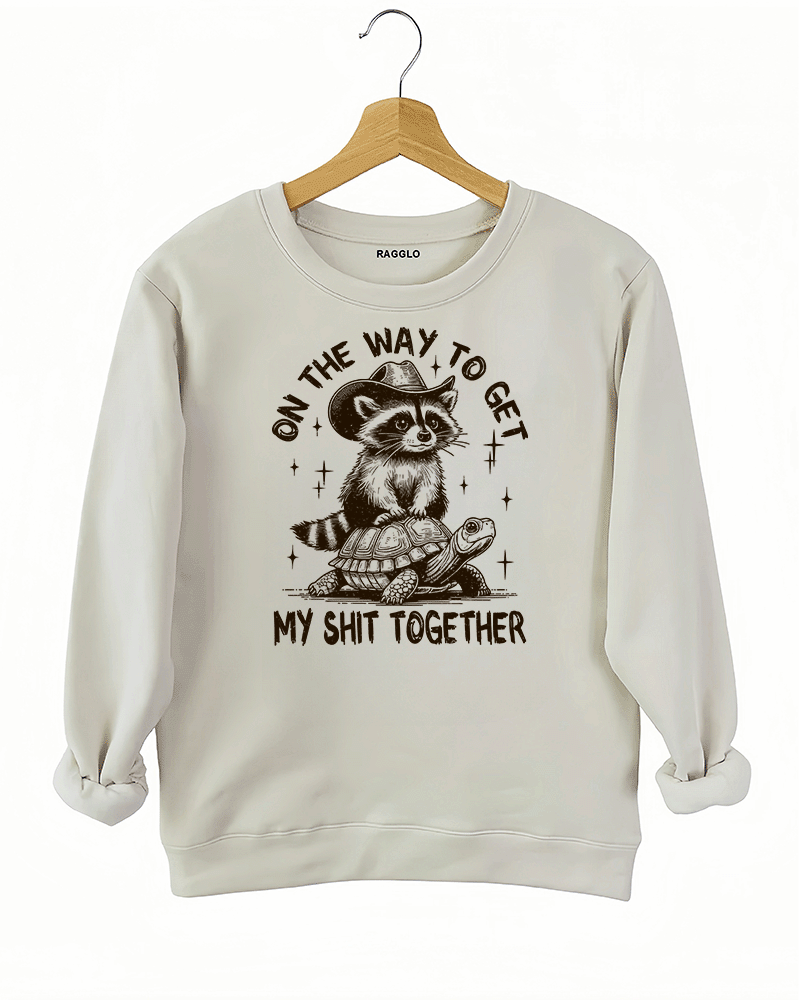 Ragglo beige sweatshirt featuring 'ON THE WAY TO GET MY SHIT TOGETHER' text with a raccoon in a cowboy hat riding a turtle, perfect for casual streetwear and humorous everyday wear.