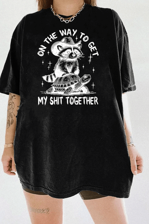 Ragglo black tee featuring 'ON THE WAY TO GET MY SHIT TOGETHER' text with a raccoon in a cowboy hat riding a turtle, perfect for casual streetwear and humorous everyday wear.