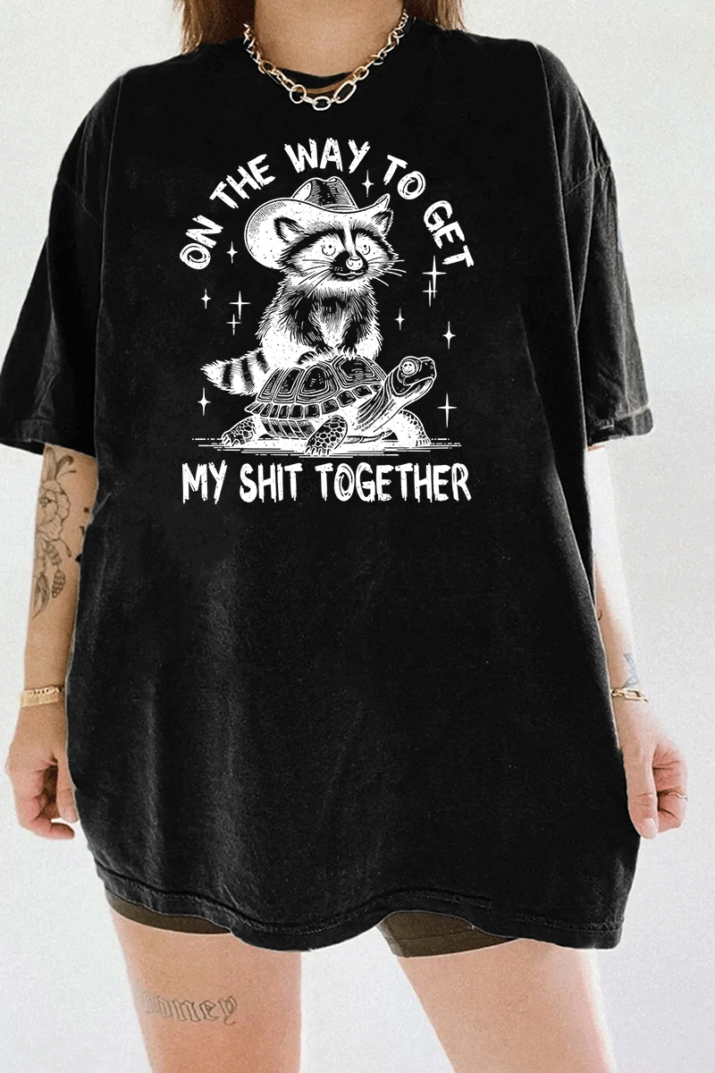 Ragglo black tee featuring 'ON THE WAY TO GET MY SHIT TOGETHER' text with a raccoon in a cowboy hat riding a turtle, perfect for casual streetwear and humorous everyday wear.