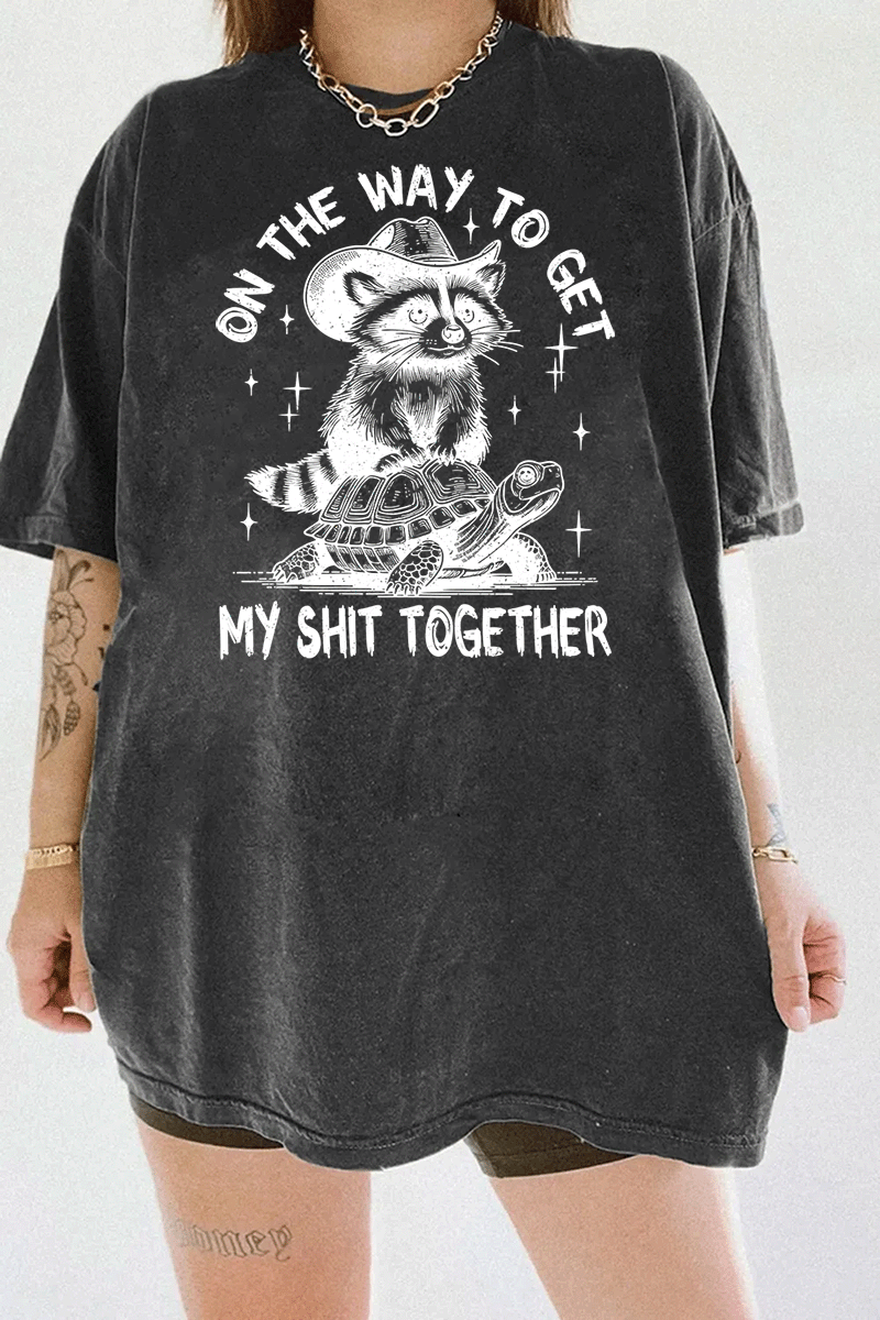 Ragglo dark gray tee featuring 'ON THE WAY TO GET MY SHIT TOGETHER' text with a raccoon in a cowboy hat riding a turtle, perfect for casual streetwear and humorous everyday wear.