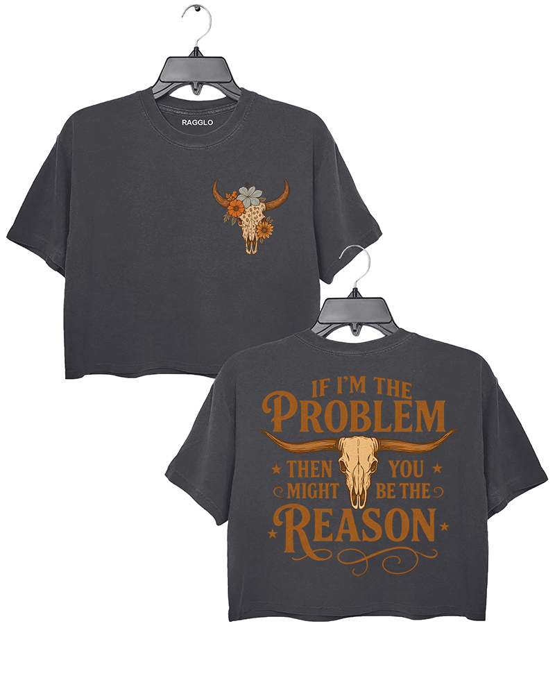Dark gray crop top with a bull skull graphic surrounded by flowers on the front, and bold text 'IF I'M THE PROBLEM THEN YOU MIGHT BE THE REASON' with a bull skull illustration on the back. Both sides feature warm orange accents.