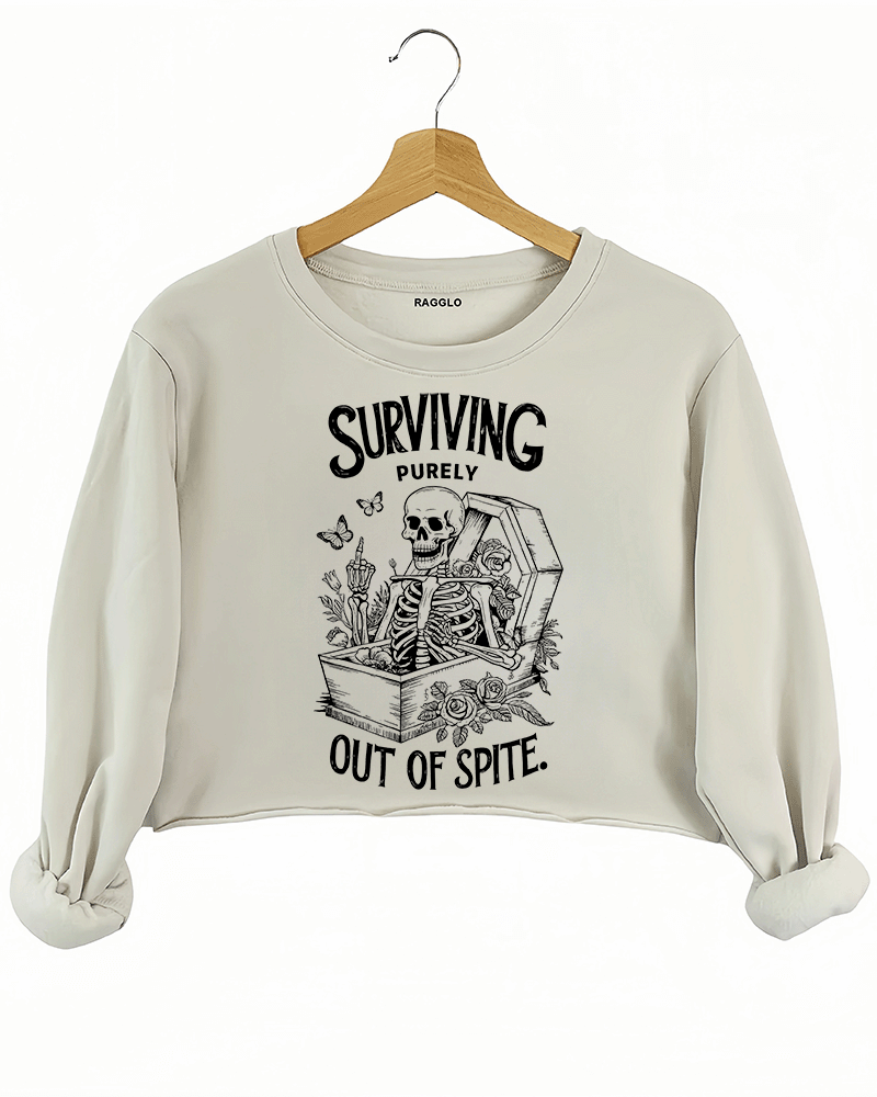 Beige crop sweatshirt with a graphic of a skeleton sitting in a coffin, adorned with roses and candles, featuring the phrase 'Surviving Purely Out of Spite,' available on Ragglo.