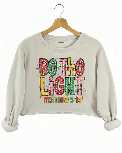 Be the Light - Matthew 5:14 graphic with polka dot letters and holiday lights on a beige crop sweatshirt, available on Ragglo.