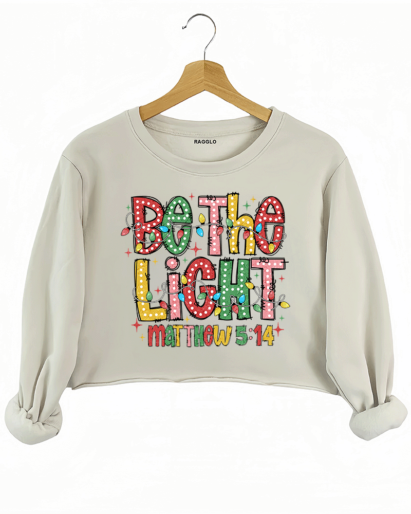 Be the Light - Matthew 5:14 graphic with polka dot letters and holiday lights on a beige crop sweatshirt, available on Ragglo.