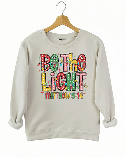 Be the Light - Matthew 5:14 graphic design with multicolored letters and festive lights on a beige sweatshirt, available on Ragglo.