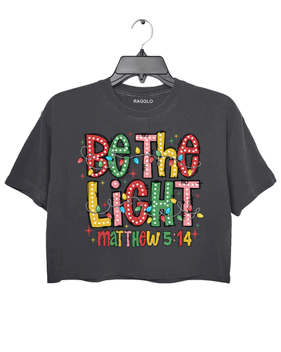 Be the Light - Matthew 5:14 graphic design featuring colorful letters and Christmas lights on a dark gray crop top, available on Ragglo.