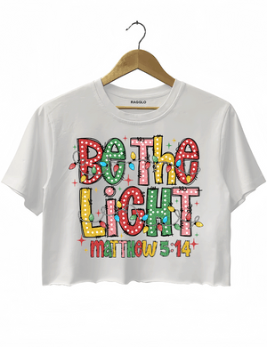 Be the Light - Matthew 5:14 graphic design with bright, colorful letters and festive lights on a white crop top, available on Ragglo.