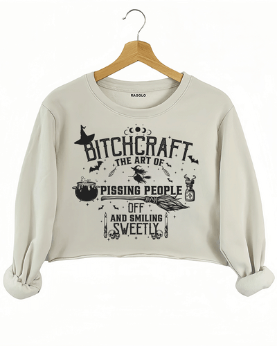 Bitchcraft design featuring the phrase 'The Art of Pissing People Off and Smiling Sweetly' along with witchcraft elements like a cauldron, broom, and skull candles, printed on a beige crop sweatshirt available at Ragglo.