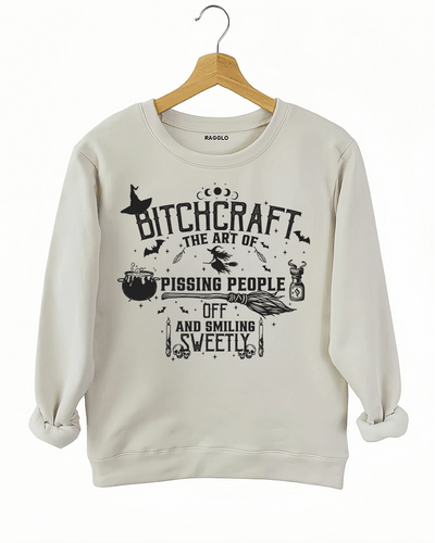 Bitchcraft graphic with the phrase 'The Art of Pissing People Off and Smiling Sweetly,' decorated with magical symbols like a witch hat, flying witch, cauldron, and potion bottle, printed on a beige sweatshirt available at Ragglo.