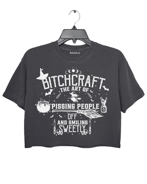 Bitchcraft graphic featuring the phrase 'The Art of Pissing People Off and Smiling Sweetly,' with witchcraft elements like a cauldron, broom, flying witch, and magic potion, printed on a dark gray crop top - available on Ragglo.