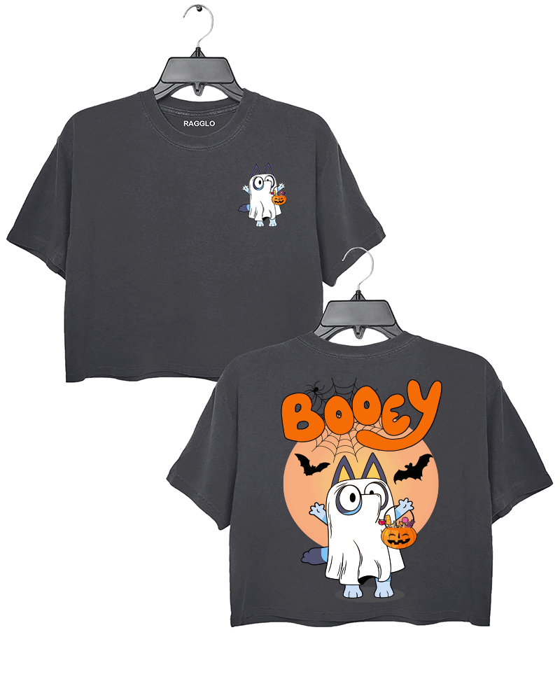 Ragglo gray crop top featuring a cute cartoon cat dressed as a ghost, holding a pumpkin-shaped candy bucket with 'BOOEY' text in bold orange letters. Halloween-themed with bats and spiderwebs in the background.