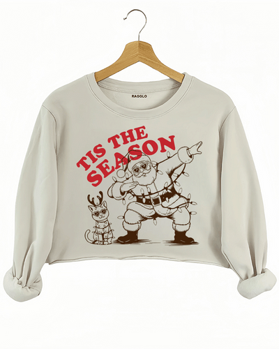 Christmas-themed Tis The Season design featuring Santa dabbing in sunglasses with a festive cat wrapped in Christmas lights, printed on a beige crop sweatshirt available at Ragglo.