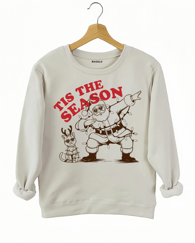 Tis The Season Christmas graphic showing Santa dabbing with a heart-shaped sunglasses and Christmas lights, along with a festive cat, printed on a beige sweatshirt available at Ragglo.