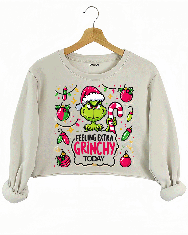 Beige crop sweatshirt featuring the iconic Grinch with a Santa hat and candy cane, set against a backdrop of holiday ornaments, available on Ragglo.