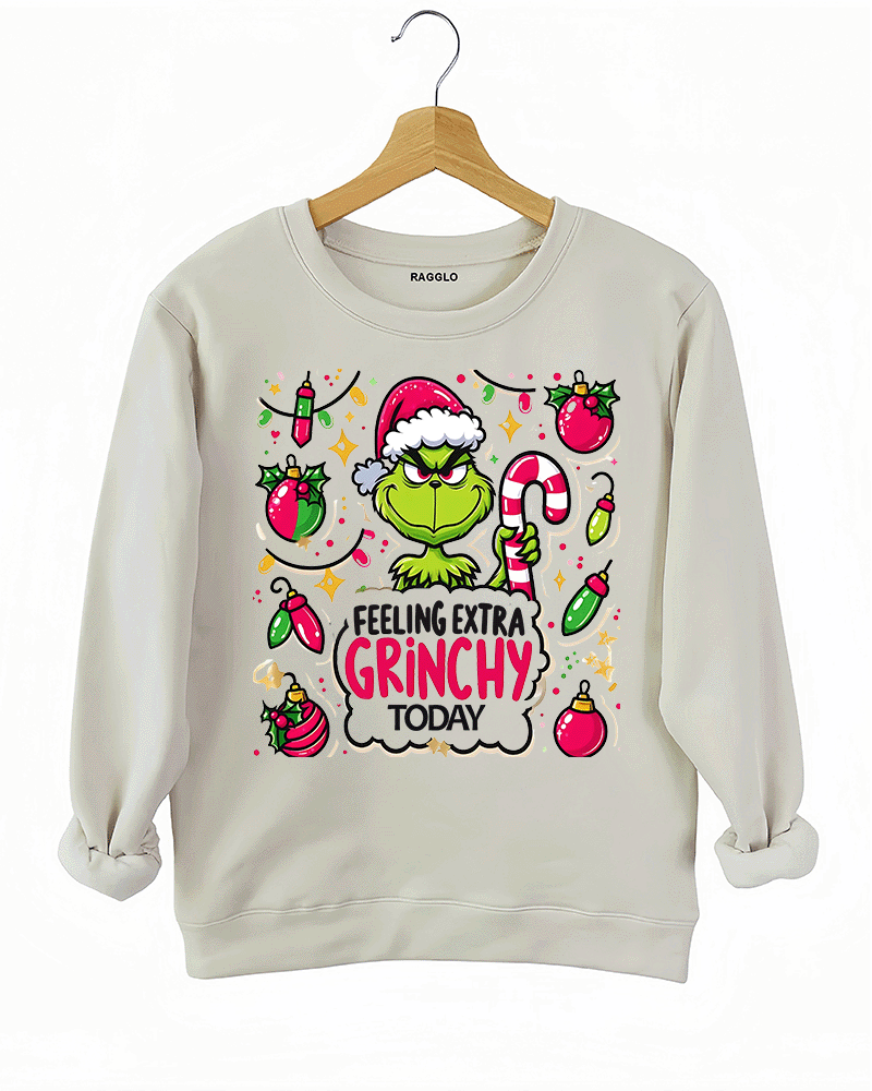 Beige sweatshirt with a festive Grinch graphic holding a candy cane, surrounded by colorful Christmas decorations, available on Ragglo.