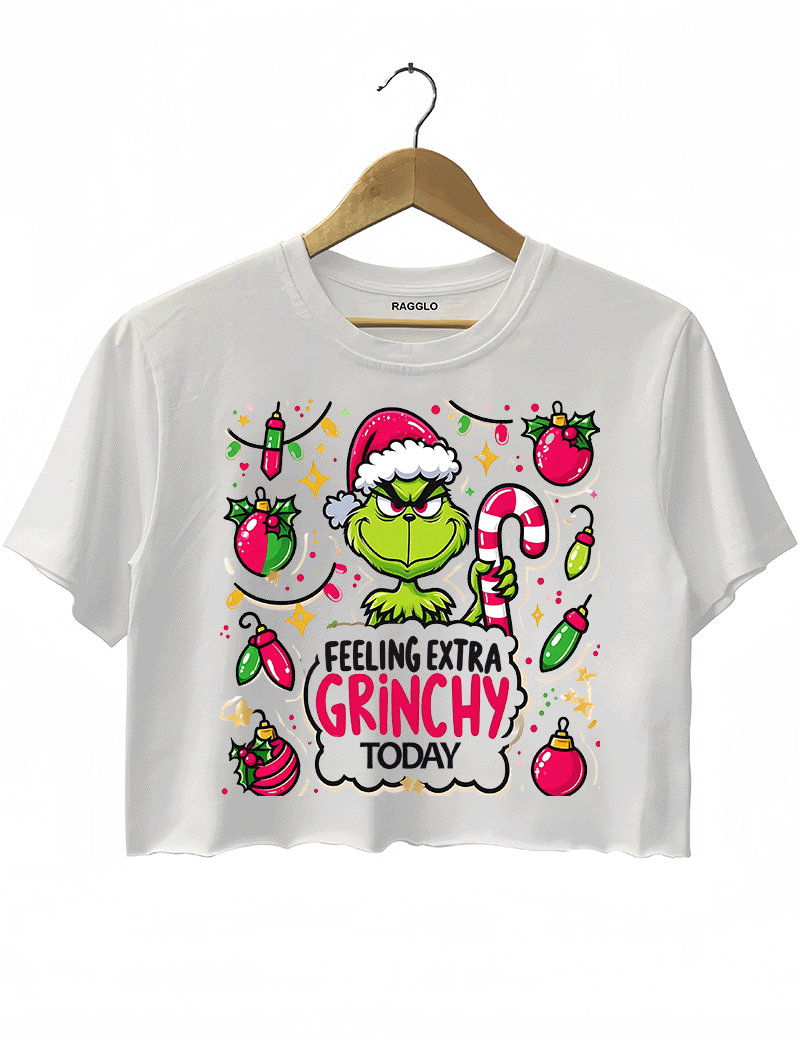 White crop top showcasing the mischievous Grinch wearing a Santa hat and holding a candy cane, with a playful Christmas ornament background, available on Ragglo.