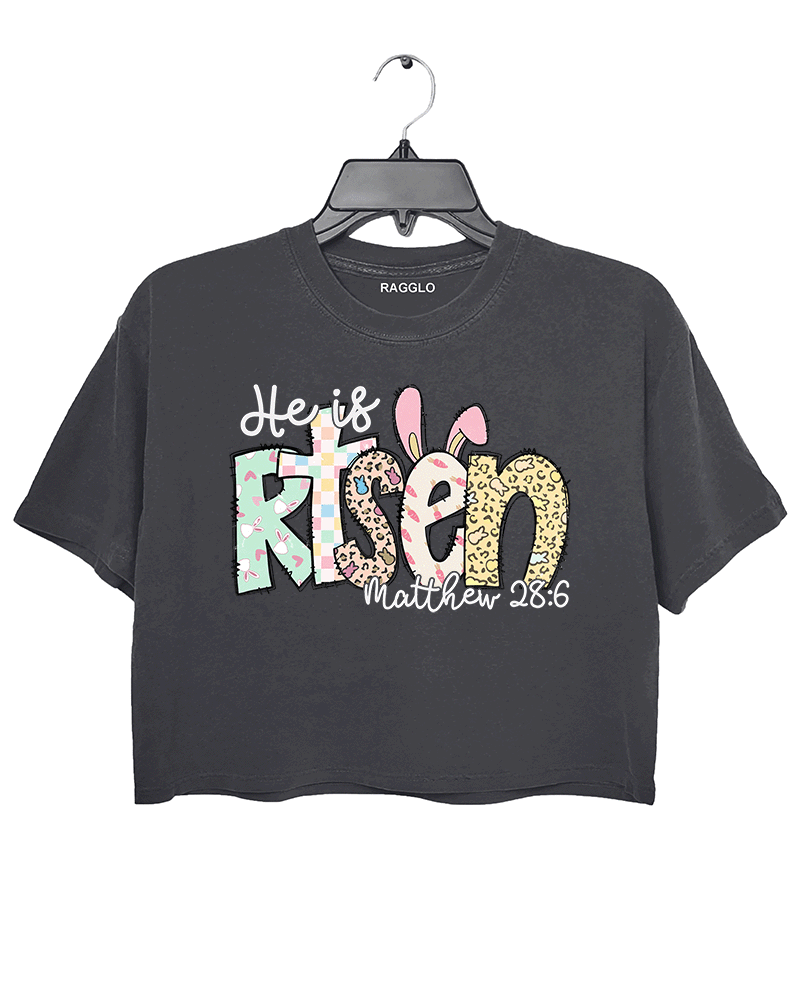Dark gray cropped women's T-shirt from Ragglo with "He is Risen" and "Matthew 28:6" text, featuring Easter-themed bunny and egg patterns in pastel colors.

