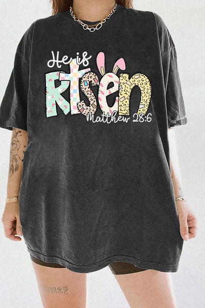 Dark gray women's T-shirt from Ragglo with "He is Risen" and "Matthew 28:6" text, featuring Easter-themed bunny and egg patterns in pastel colors.

