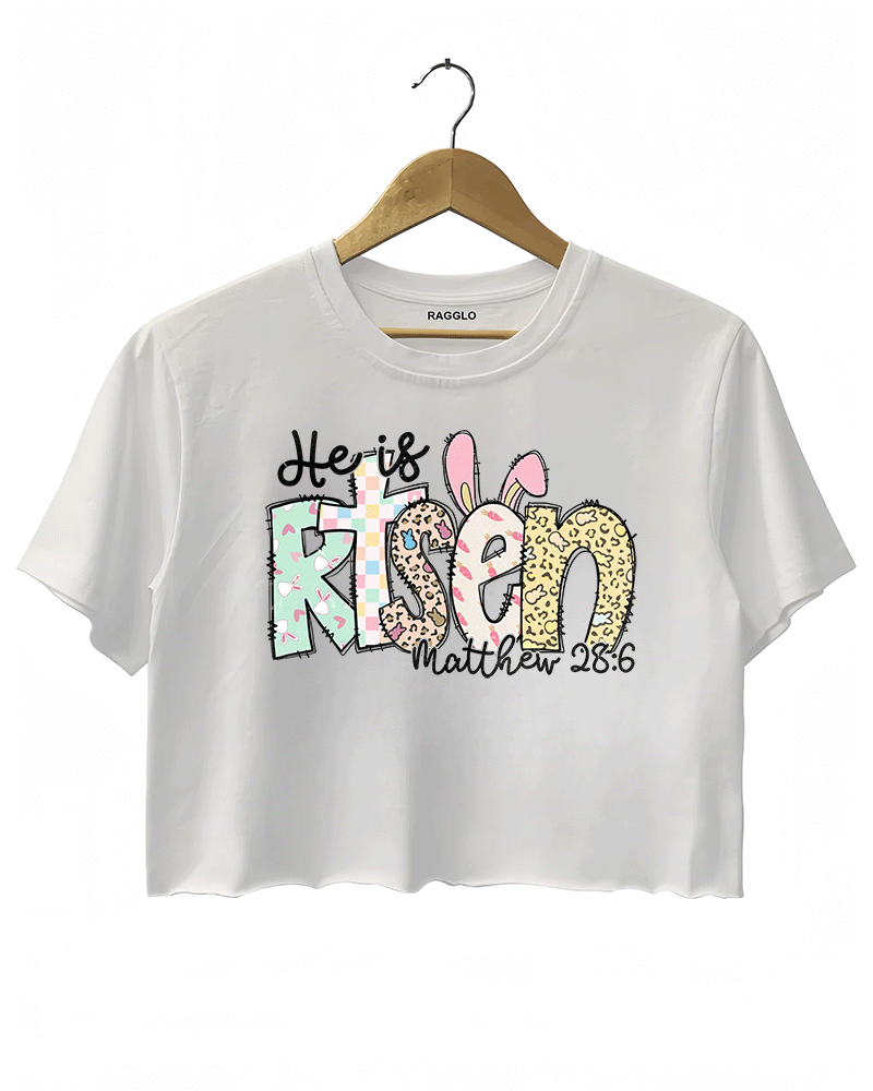 White cropped women's T-shirt from Ragglo with "He is Risen" and "Matthew 28:6" text, featuring Easter-themed bunny and egg patterns in pastel colors.

