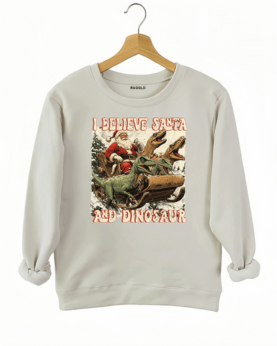 Whimsical graphic of Santa Claus in a sleigh pulled by dinosaurs with the text 'I Believe in Santa and Dinosaur' on a beige sweatshirt, offered by Ragglo.