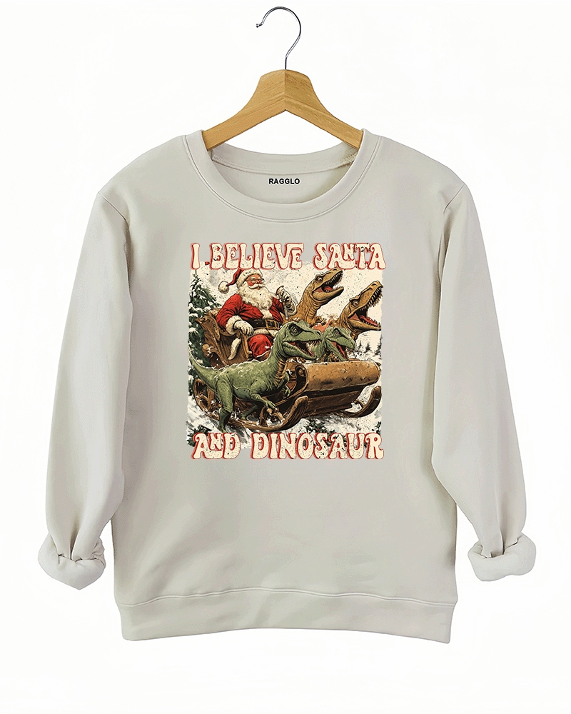 Whimsical graphic of Santa Claus in a sleigh pulled by dinosaurs with the text 'I Believe in Santa and Dinosaur' on a beige sweatshirt, offered by Ragglo.