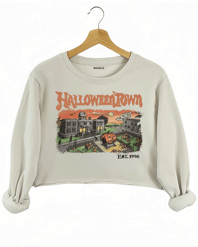 Ragglo beige women's crop sweatshirt featuring a spooky town with a jack-o'-lantern, graveyard, and old houses under a sunset sky with bats, and the text 'Halloween Town Est. 1998' in a retro Halloween graphic design.