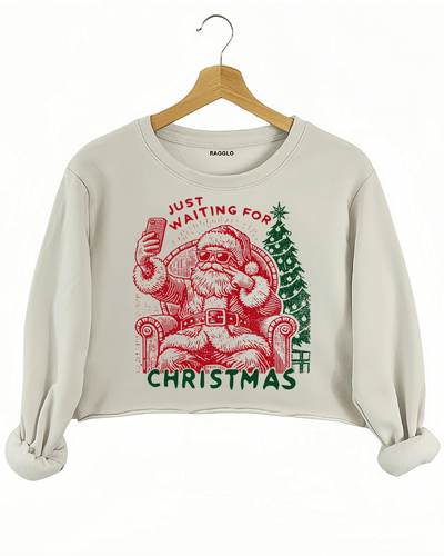 Ragglo beige crop sweatshirt featuring a Santa selfie graphic with sunglasses, a Christmas tree, and 'JUST WAITING FOR CHRISTMAS' text, perfect for festive holiday streetwear and fun celebrations.

