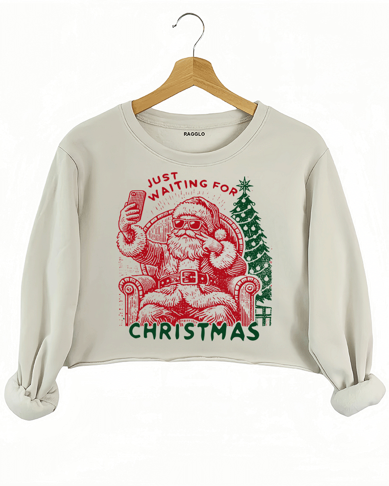 Ragglo beige crop sweatshirt featuring a Santa selfie graphic with sunglasses, a Christmas tree, and 'JUST WAITING FOR CHRISTMAS' text, perfect for festive holiday streetwear and fun celebrations.

