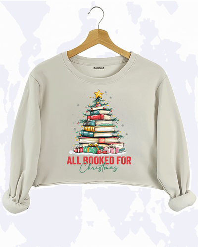 Ragglo beige crop sweatshirt featuring a Christmas tree made of stacked books, decorated with lights, stars, and gifts, with 'ALL BOOKED FOR CHRISTMAS' text, perfect for festive streetwear for book lovers.

