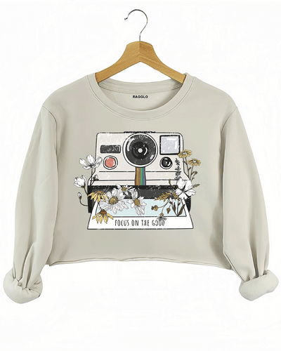 Ragglo beige crop sweatshirt featuring a vintage Polaroid camera surrounded by flowers, with 'FOCUS ON THE GOOD' text, perfect for retro-inspired streetwear with a positive vibe.

