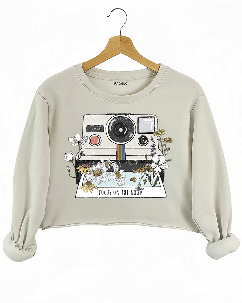 Ragglo beige crop sweatshirt featuring a vintage Polaroid camera surrounded by flowers, with 'FOCUS ON THE GOOD' text, perfect for retro-inspired streetwear with a positive vibe.

