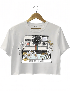 Ragglo white crop top featuring a vintage Polaroid camera surrounded by flowers, with 'FOCUS ON THE GOOD' text, perfect for retro-inspired streetwear with a positive vibe.


