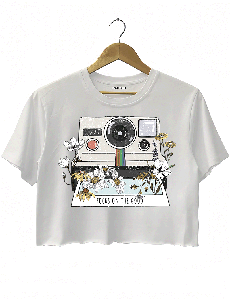 Ragglo white crop top featuring a vintage Polaroid camera surrounded by flowers, with 'FOCUS ON THE GOOD' text, perfect for retro-inspired streetwear with a positive vibe.

