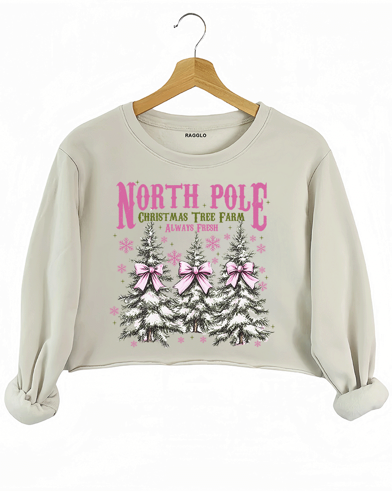 Ragglo beige crop sweatshirt featuring three Christmas trees with pink bows and snowflakes, with 'NORTH POLE CHRISTMAS TREE FARM ALWAYS FRESH' text, perfect for festive holiday streetwear.


