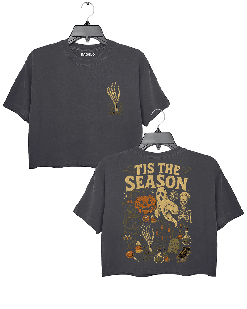 Dark gray cropped T-shirt by RAGGLO with spooky Halloween graphics on both front and back. Front features a gold skeleton hand reaching up with a small flame. Back showcases a bold vintage-style Halloween scene with a pumpkin head, ghost, skeleton, potion bottles, candy, and the phrase "'TIS THE SEASON" in large gold lettering. A perfect mix of eerie and retro charm.