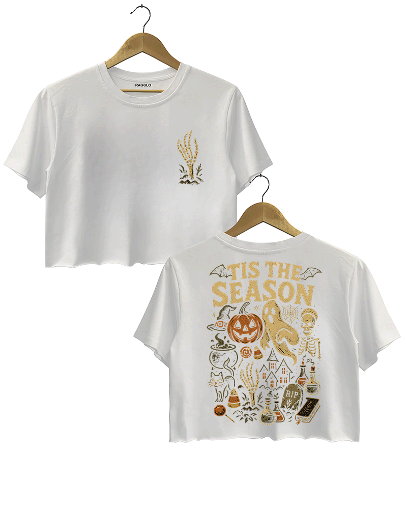 White cropped T-shirt by RAGGLO with Halloween-themed graphics on both the front and back. The front features a subtle gold skeleton hand reaching upwards, holding a flame. The back showcases a vintage Halloween scene with a pumpkin, ghost, skeleton, potion bottles, candy, and the phrase "'TIS THE SEASON" in large golden lettering. A bold and festive design perfect for the spooky season.