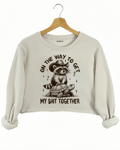 Humorous design of a raccoon in a cowboy hat riding a turtle with text 'On the Way to Get My Shit Together' on a beige crop sweatshirt, available on Ragglo.