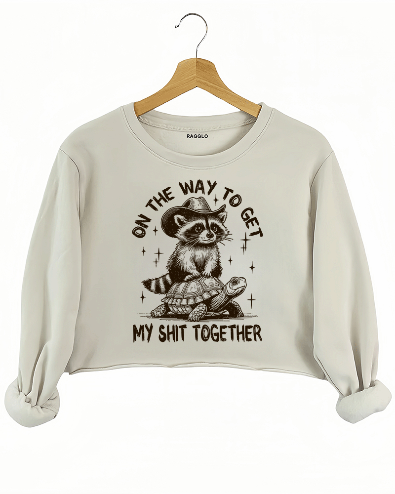 Humorous design of a raccoon in a cowboy hat riding a turtle with text 'On the Way to Get My Shit Together' on a beige crop sweatshirt, available on Ragglo.